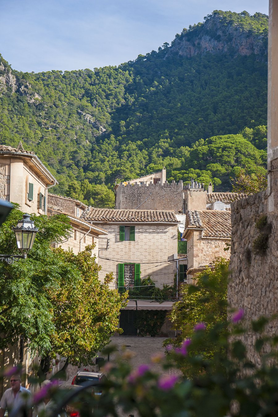 Calle de un pueblo español con casas de piedra y contraventanas verdes junto a una montaña boscosa